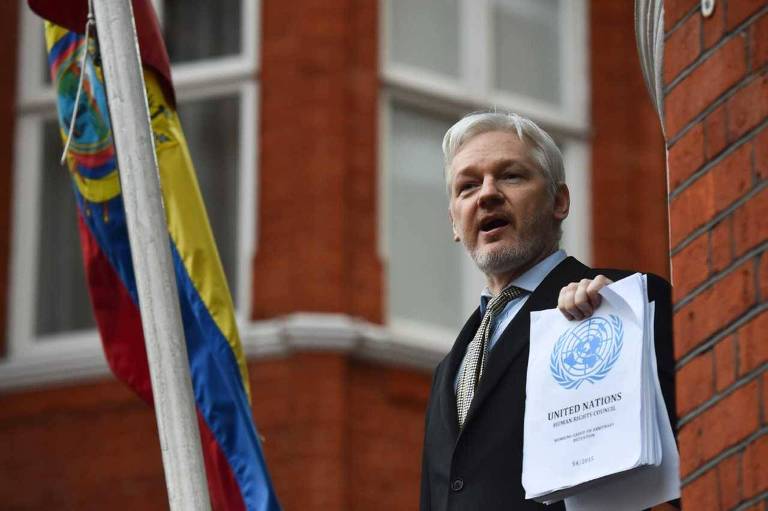 Um homem de cabelos grisalhos e barba curta está segurando um documento com o logotipo das Nações Unidas. Ele está em frente a um prédio de tijolos vermelhos da embaixada do Equador, ao lado de uma bandeira do Equador.