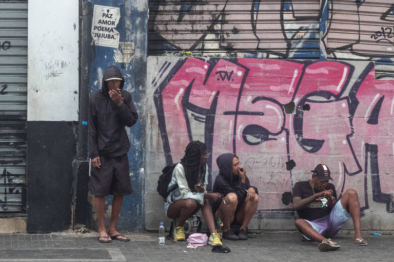 A imagem mostra um grupo de quatro pessoas sentadas em um calçadão, com uma parede grafitada ao fundo. Uma das pessoas está em pé, fumando, enquanto as outras três estão sentadas no chão, algumas com mochilas e um chapéu. A parede atrás deles possui uma grande arte de grafite em tons de rosa e roxo
