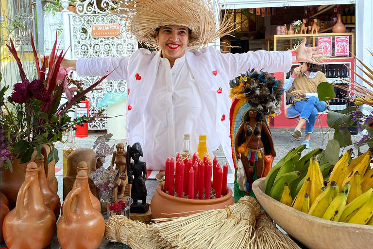 Uma mulher vestida de branco com chapéu de palha abre os braços de frente a uma mesa com objetos de artesanato