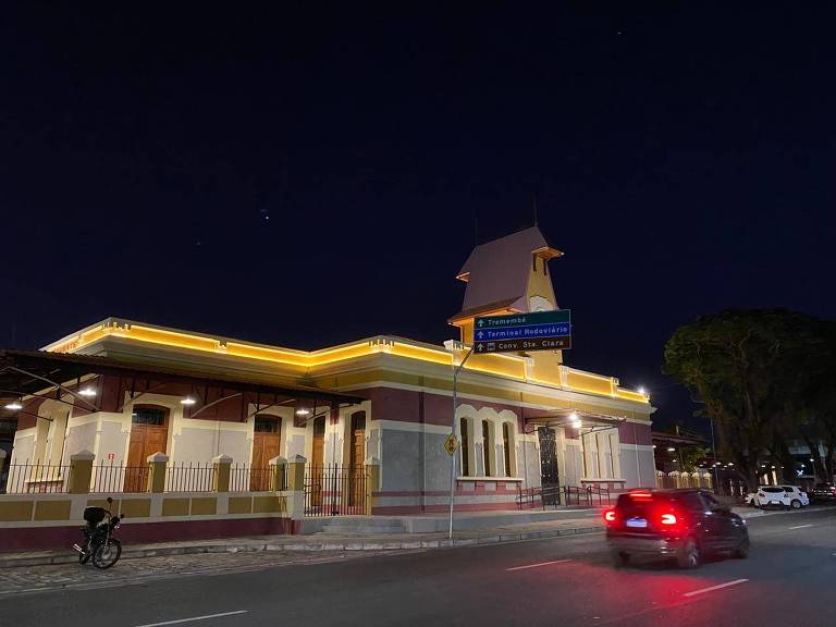 Imagem noturna mostra a estação ferroviária de Taubaté iluminada, com um veículo passando à sua frente