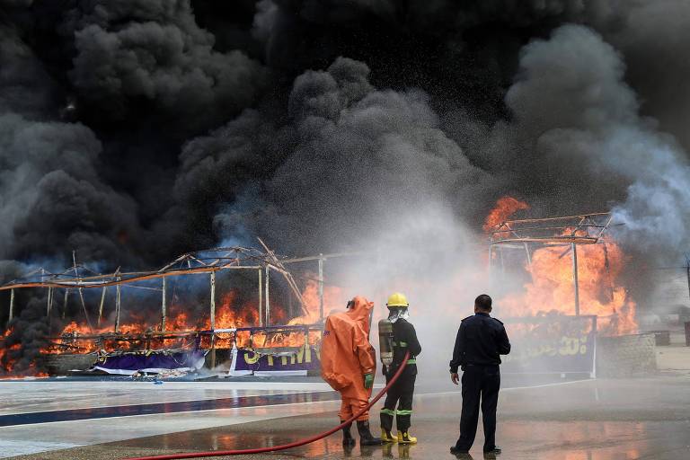 A imagem mostra um grande incêndio em uma estrutura ao ar livre, com chamas intensas e fumaça preta densa. Três bombeiros, dois deles usando uniformes de proteção e capacetes, estão no primeiro plano tentando controlar o fogo com mangueiras de água.