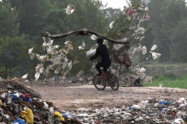 Garoto anda de bicicleta entre três pilhas de sacolas plásticas descartadas em um lixão nos arredores de Laore, no Paquistão
