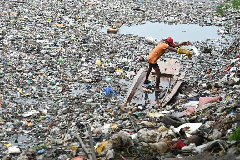 Trabalhador de limpeza urbana abre caminho em meio ao lixo plástico que boia em um canal de Chennai, na Índia
