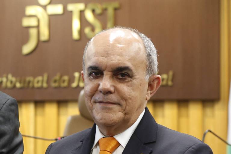 O ministro está vestido com terno e gravata laranja; ao fundo a inscrição TST