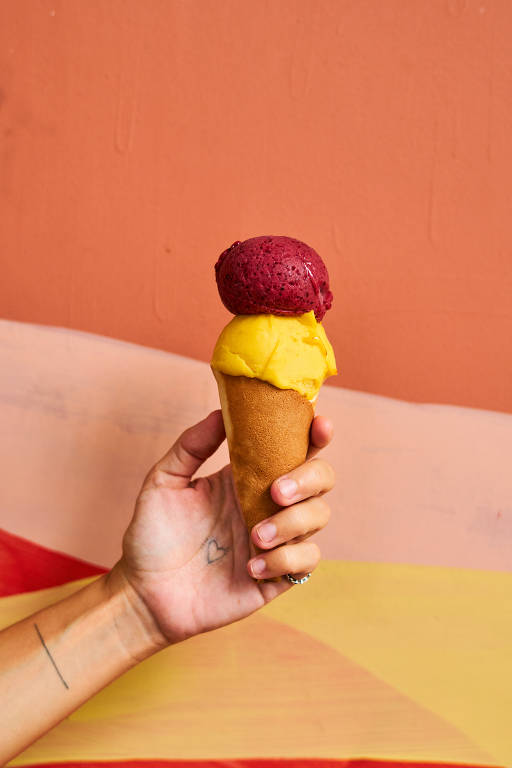 A imagem mostra uma mão segurando um sorvete de casquinha com duas bolas. A bola superior é de cor roxa, enquanto a bola inferior é amarela. A mão que segura o sorvete tem uma tatuagem de coração no pulso e uma linha reta no antebraço. O fundo é uma parede com tons de laranja e rosa.