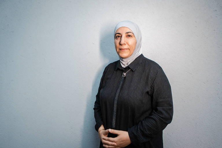 Mulher usando um hijab branco e uma roupa preta, posando contra uma parede cinza. Ela está olhando diretamente para a câmera com uma expressão neutra e suas mãos estão cruzadas na frente do corpo.