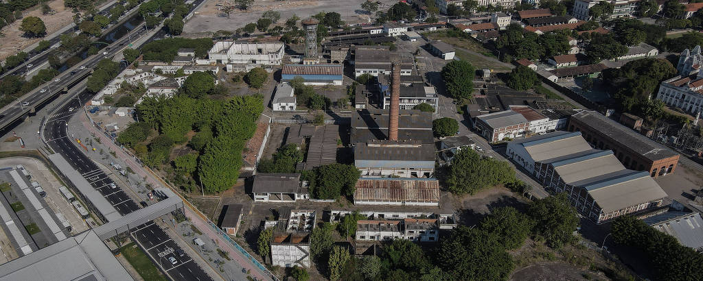 Imagem aérea de um complexo industrial cercado por vegetação e várias estradas. O complexo possui várias construções, incluindo uma alta chaminé de tijolos. Ao redor, há áreas urbanas com edifícios e estradas movimentadas.