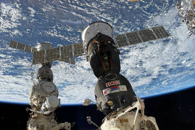 Imagem no espaço mostra nave com dois painéis solares abertos acoplada a uma estrutura, com a Terra ao fundo
