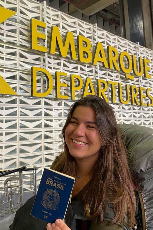 Uma mulher sorridente está no aeroporto, em frente a um grande letreiro amarelo que diz 'EMBARQUE DEPARTURES'. Ela segura um passaporte brasileiro e carrega uma mochila grande nas costas. Ao fundo, há carrinhos de bagagem e uma parede com um padrão geométrico.