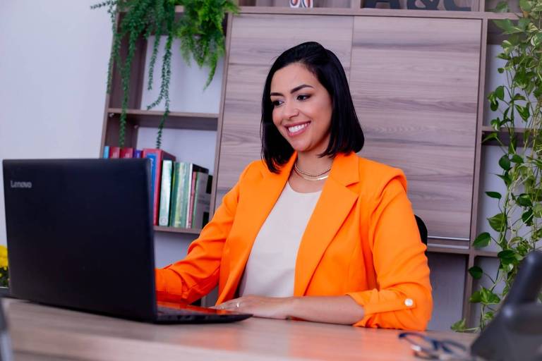 Amanda, uma mulher com cabelos pretos e lisos na altura do ombro, está sentada em uma mesa, trabalhando em um laptop. Ela está sorrindo e vestindo um blazer laranja sobre uma blusa branca. Ao fundo, há uma estante com livros e plantas decorativas.