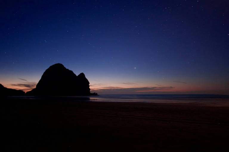 A imagem mostra uma praia ao anoitecer, com um grande rochedo escuro em silhueta contra o céu. O céu está em tons de azul escuro e laranja, com várias estrelas visíveis. O mar está calmo e reflete as cores do céu.