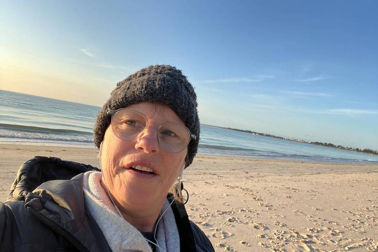 Uma mulher usando óculos, gorro de lã cinza e casaco preto está tirando uma selfie na praia. O mar está calmo ao fundo, e o céu está claro com poucas nuvens. A areia da praia está marcada com pegadas.