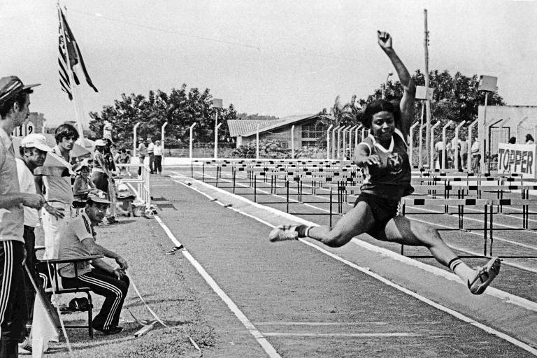 A imagem em preto e branco mostra uma atleta no meio de um salto em distância em uma pista de atletismo. A atleta está no ar, com uma perna estendida para a frente e a outra para trás. À esquerda, há várias pessoas observando, algumas sentadas e outras em pé. Ao fundo, é possível ver uma série de barreiras de corrida e algumas construções.
