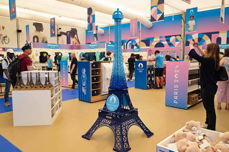 A imagem mostra uma loja de souvenirs com tema de Paris. No centro, há uma réplica azul da Torre Eiffel. Ao redor, há várias prateleiras e expositores com produtos diversos, incluindo roupas, pelúcias e outros itens. As paredes e o teto têm uma decoração colorida, predominantemente em tons de azul e rosa. Há várias pessoas na loja, algumas olhando os produtos e outras tirando fotos.
