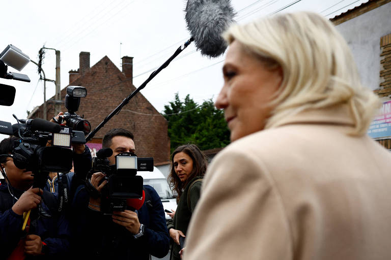 Marine Le Pen, mulher de cabelos loiros, está sendo entrevistada ao ar livre por um grupo de jornalistas. Ela está de perfil, vestindo um casaco bege. Os jornalistas estão segurando câmeras e microfones, e há um microfone boom visível acima da mulher. Ao fundo, há uma casa de tijolos e algumas árvores.