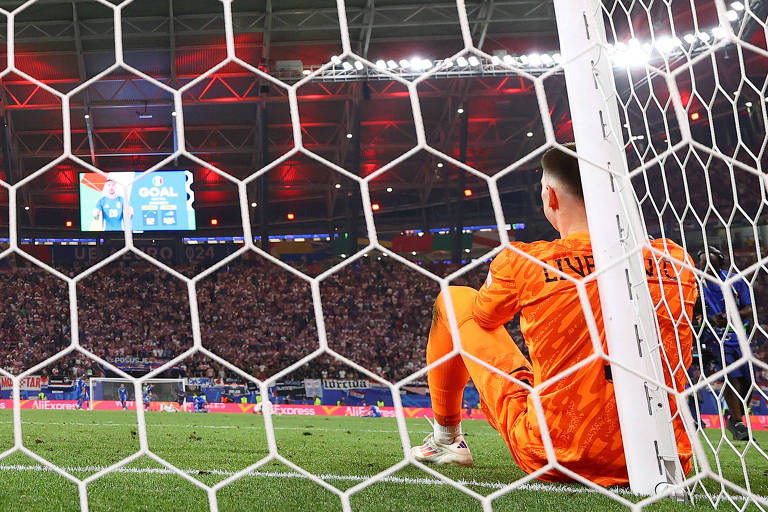 O goleiro croata Livakovic, vestindo uniforme laranja, sentado e desolado encostado na trave de seu gol, enquanto o placar, ao fundo, mostra o gol da Itália, feito por Zaccagni nos acréscimos do 2º tempo na Eurocopa na Alemanha