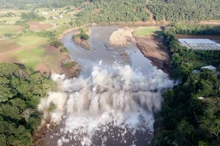Imagem aérea de uma explosão controlada em uma barragem de um rio, com uma grande quantidade de fumaça e água sendo lançada para o ar. A área ao redor é cercada por vegetação densa e algumas construções. O rio segue seu curso natural, com áreas de terra visíveis nas margens.