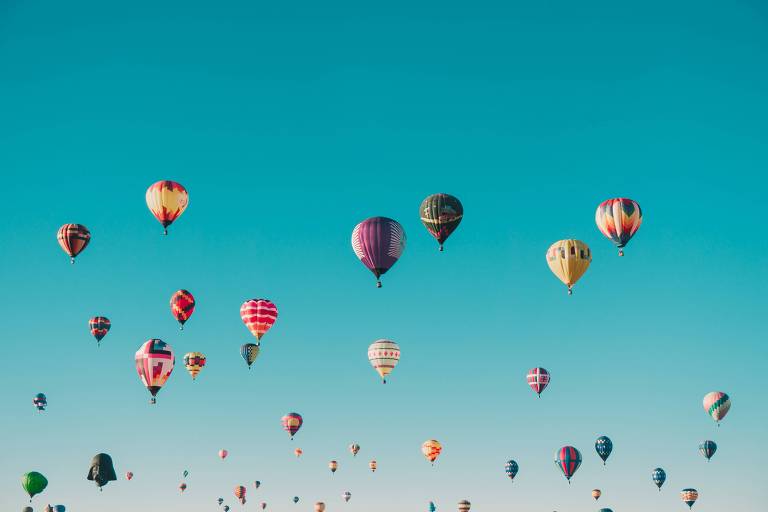 A imagem mostra vários balões de ar quente coloridos flutuando no céu azul claro. Os balões têm diferentes padrões e cores, incluindo listras, xadrez e formas geométricas. O céu está limpo, sem nuvens, proporcionando um fundo claro e uniforme para os balões.