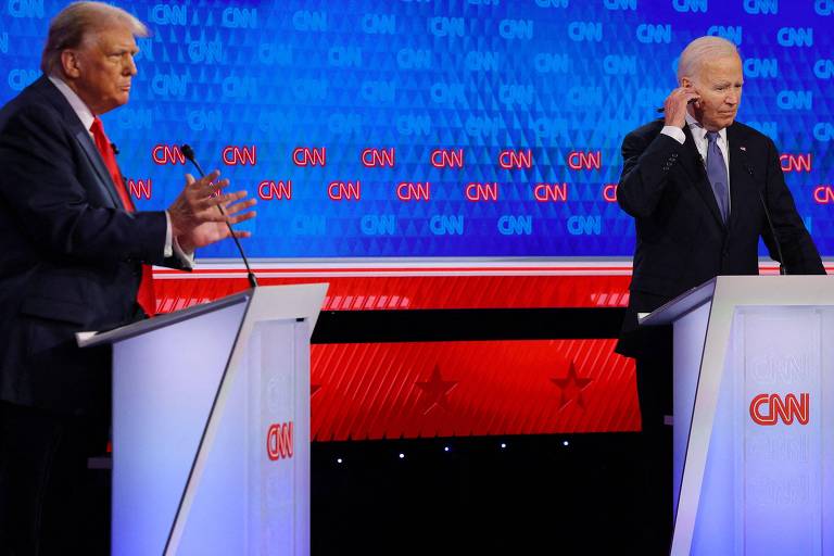 A imagem mostra dois homens em pé atrás de púlpitos durante um debate presidencial transmitido pela CNN. O homem à esquerda está gesticulando com as mãos, enquanto o homem à direita está tocando sua orelha. O fundo é azul com o logotipo da CNN repetido várias vezes.