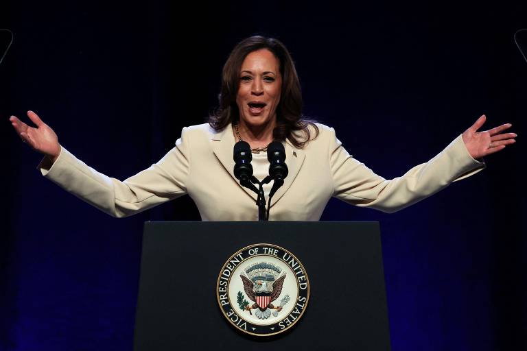 Uma pessoa está discursando em um pódio com o selo do Vice-Presidente dos Estados Unidos. Ela está usando um blazer claro e tem os braços abertos. O fundo é escuro e há dois microfones no pódio.