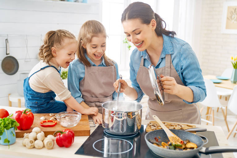 A imagem mostra uma mulher e duas crianças cozinhando juntas em uma cozinha bem iluminada. A mulher está mexendo numa panela no fogão enquanto as crianças observam com entusiasmo. Há vários ingredientes frescos, como pimentões, cogumelos e tomates, sobre a bancada. Uma frigideira com legumes também está no fogão.
