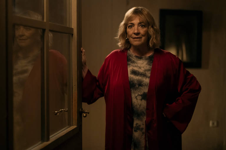 Uma mulher de cabelos loiros está em pé na porta de uma sala, segurando a maçaneta com uma mão. Ela veste um robe vermelho sobre uma camiseta estampada. A sala está iluminada de forma suave e há um espelho na parede ao fundo, refletindo parcialmente a mulher