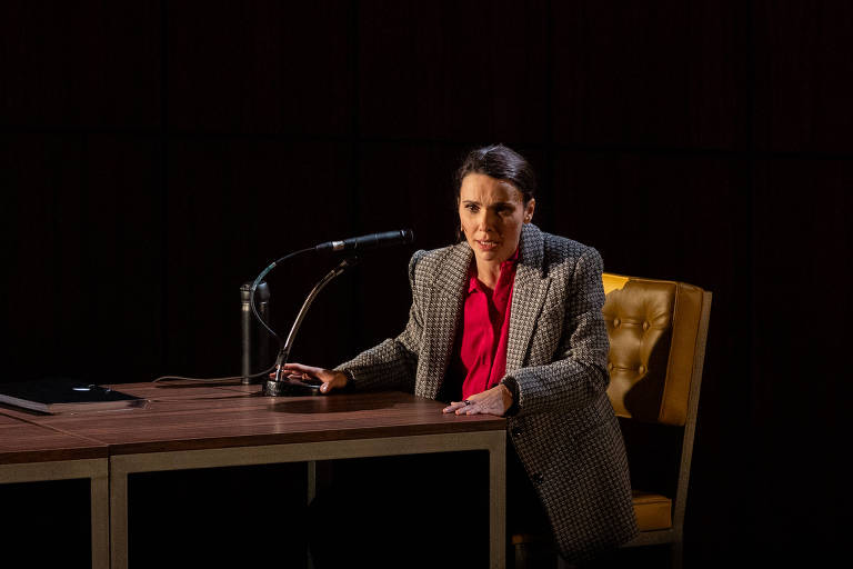Uma mulher está sentada em uma cadeira, vestindo um blazer cinza e uma camisa vermelha. Ela está em uma mesa de madeira com um microfone à sua frente. A expressão dela parece séria e concentrada. O fundo é escuro.
