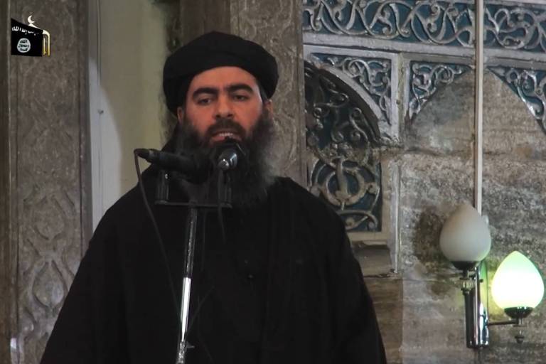 Um homem com barba e vestindo roupas pretas está falando ao microfone. Ele usa um turbante preto e está em frente a uma parede decorada com padrões intrincados. Há uma lâmpada acesa ao lado direito da imagem. No canto superior esquerdo, há um logotipo com texto em árabe.