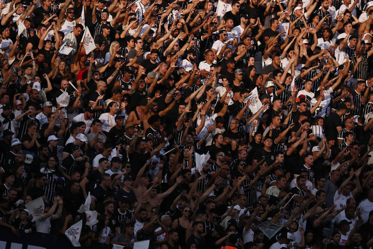 Uma multidão de torcedores do Corinthians grita, com os braços esticados para a frente e o alto