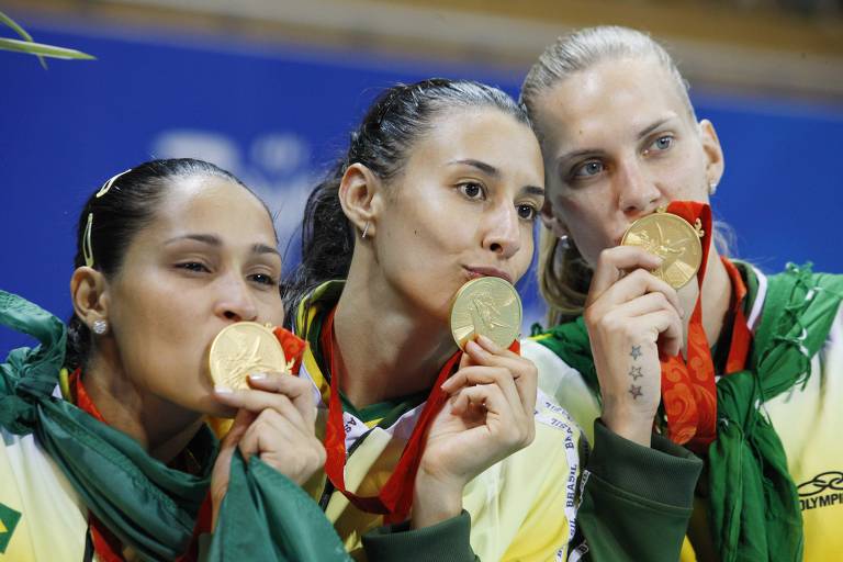 Três atletas femininas estão segurando e beijando suas medalhas de ouro. Elas estão vestindo uniformes com as cores verde e amarelo, e têm medalhas penduradas em fitas vermelhas ao redor do pescoço. As atletas parecem estar celebrando uma vitória.