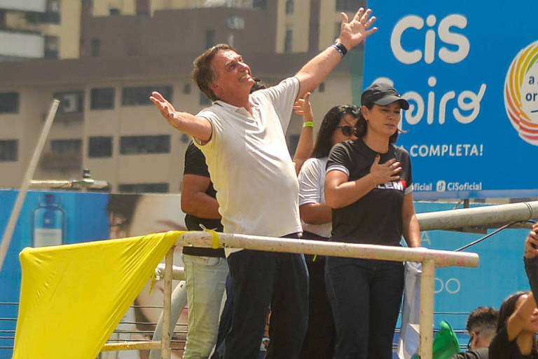 Imagem mostra Jair Bolsonaro, homem de camiseta branca com os braços abertos, em um evento ao ar livre. Ao fundo, há outras pessoas e um banner azul com o texto 'CIS' e 'completa'.