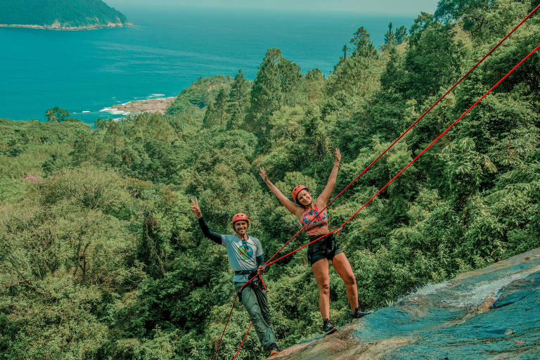 Casal preso por uma corda de rapel desce penhasco de cachoeira com 90 metros de altura. Ao fundo há um trecho de mata antes do mar