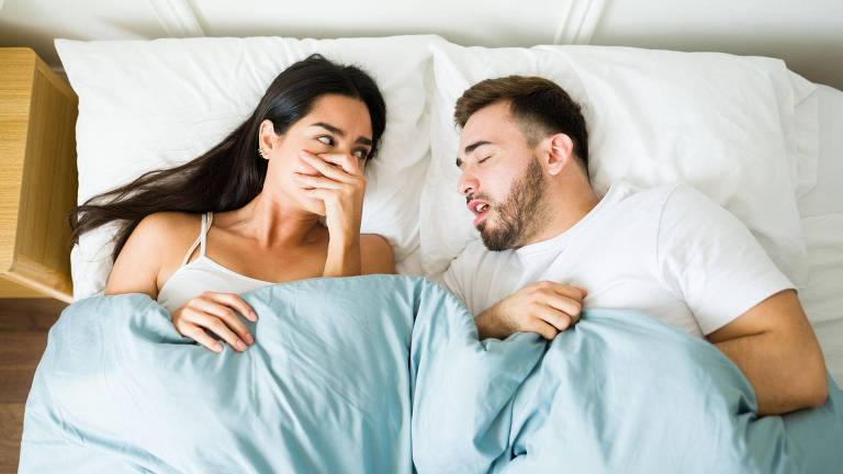Casal deitado na cama, mulher tapando o nariz enquanto o homem dorme de boca aberta