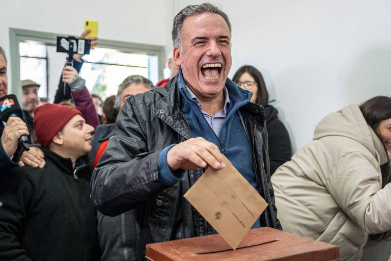 Um homem sorridente está depositando um envelope em uma urna de votação de madeira. Ele veste uma jaqueta preta e uma camisa azul. Ao fundo, várias pessoas observam a cena, algumas tirando fotos e outras conversando. Uma pessoa está usando um gorro vermelho e outra está com um casaco bege.
