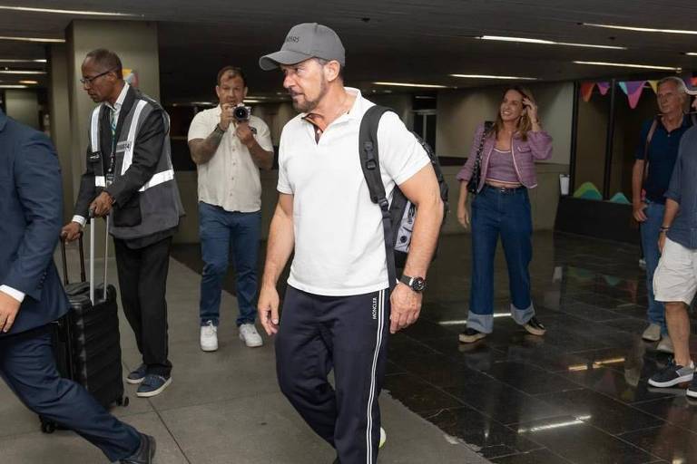 Homem de barba, camisa branca e boné anda em aeroporto brasileiro