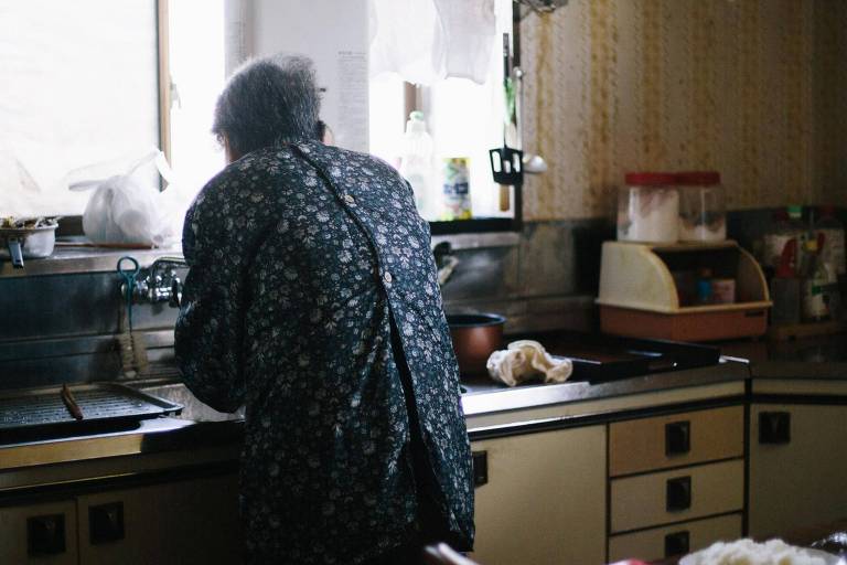 A imagem mostra uma pessoa idosa de costas, usando um casaco estampado, lavando louça em uma pia de cozinha. A cozinha tem um estilo antigo, com armários de cor clara e puxadores escuros. Há diversos itens sobre a pia, incluindo uma garrafa plástica e um pano de prato.