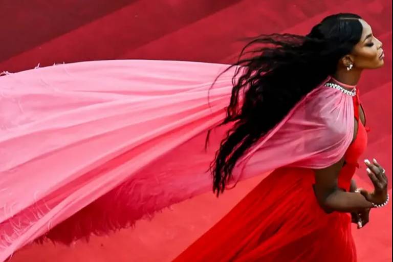 Imagem de uma mulher usando um vestido vermelho longo com uma capa rosa esvoaçante. Ela está em um ambiente com um tapete vermelho e tem cabelos longos e soltos. A capa rosa tem detalhes de plumas na borda.