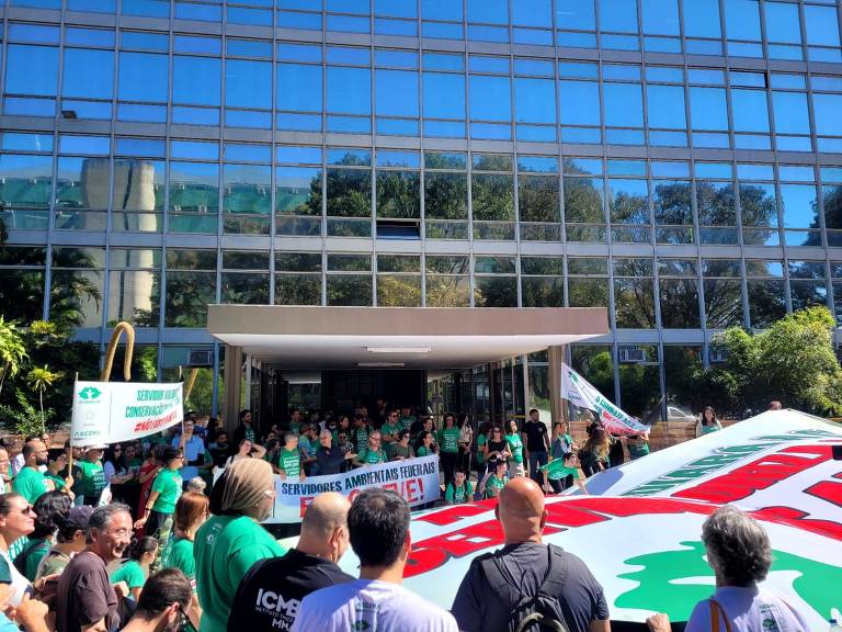 Pessoas com faixas e vestindo camisetas verdes fazem manifestação em frente a prédio espelhado