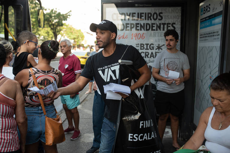 Fotografia de Rick Azevedo distribuindo panfletos para pessoas em um ponto de ônibus. Ele está vestindo uma camiseta preta e um boné preto, ambos com a inscrição 'VAT'. Envolta, pessoas aguardam para embarcar no ônibus.