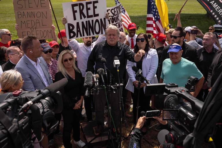 A imagem mostra um grupo de pessoas em um protesto ao ar livre. No centro, um homem está falando em um microfone, cercado por várias câmeras e microfones de imprensa. Algumas pessoas seguram cartazes, incluindo um que diz 'FREE BANNON' e outro que diz 'STEVE BANNON MEDAL OF FREEDOM'. Há também várias bandeiras americanas visíveis no fundo.