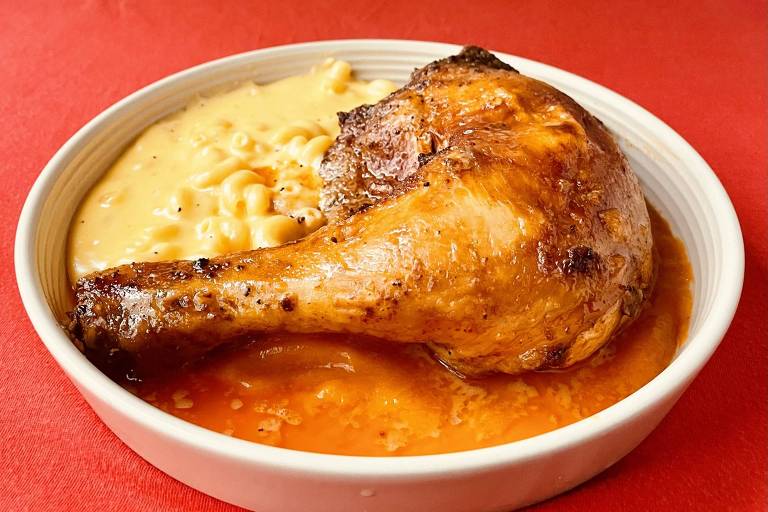 Imagem de um prato branco contendo uma coxa de frango assado com pele dourada e crocante, acompanhada de macarrão com queijo cremoso. O prato está sobre uma superfície vermelha.