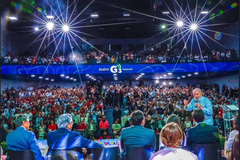 No primeiro plano, pessoas sentadas em cadeiras de costas para a foto. Depois, está Lula, homem branco de cabelo grisalho e camisa azul, discursa com microfone na mão, em pé. Ao fundo, plateia em dois pisos, em local fechado, com luzes refletindo ao fundo, no alto.