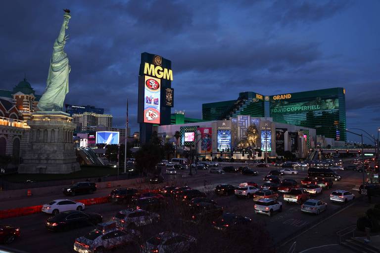 A imagem mostra uma vista noturna da Las Vegas Strip, com destaque para o hotel e cassino MGM Grand, iluminado com luzes verdes. À esquerda, há uma réplica da Estátua da Liberdade. A rua está movimentada com muitos carros. Placas de neon e letreiros iluminados são visíveis, incluindo um que menciona 'David Copperfield'