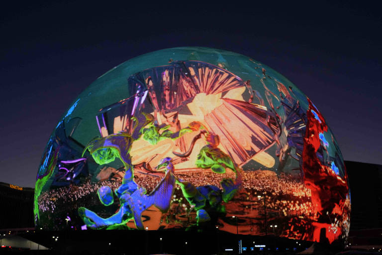 A imagem mostra uma grande cúpula iluminada à noite com uma exibição de luzes coloridas e padrões abstratos. As cores predominantes são verde, azul, vermelho e laranja, criando um efeito visual vibrante e dinâmico. O fundo é um céu noturno escuro, destacando ainda mais as luzes da cúpula.