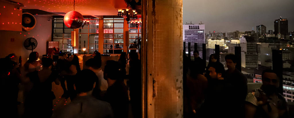 A imagem mostra uma festa em um terraço à noite. À esquerda, há uma área interna com luzes vermelhas e várias pessoas reunidas. À direita, há uma área externa com uma vista da cidade ao fundo, iluminada por luzes urbanas. Uma luminária de parede está acesa, iluminando a área externa.