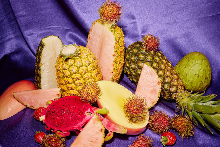 A imagem mostra uma variedade de frutas tropicais dispostas sobre um fundo de tecido roxo. Entre as frutas, há abacaxis inteiros e cortados, uma pitaya cortada ao meio, uma manga cortada ao meio, morangos, rambutãs e uma fruta verde que parece ser uma cherimóia.