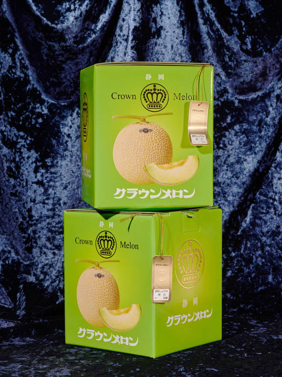 A imagem mostra duas caixas verdes empilhadas, ambas com a imagem de um melão e uma fatia de melão na frente. As caixas têm o texto 'Crown Melon' e caracteres japoneses. Há também uma etiqueta dourada pendurada em cada caixa. O fundo é um tecido azul escuro com textura.