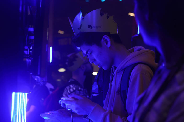 A imagem mostra um jovem usando uma coroa de papel e um moletom com capuz, concentrado em um dispositivo eletrônico em suas mãos. Ele está em um ambiente escuro, iluminado por luzes azuis. Outras pessoas e elementos do ambiente também são visíveis, mas estão desfocados.