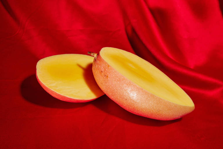 A imagem mostra uma manga cortada ao meio, com uma das metades deitada e a outra em pé, sobre um tecido vermelho. A polpa da manga é amarela e a casca é rosada com algumas manchas escuras. O fundo vermelho realça as cores vibrantes da fruta.