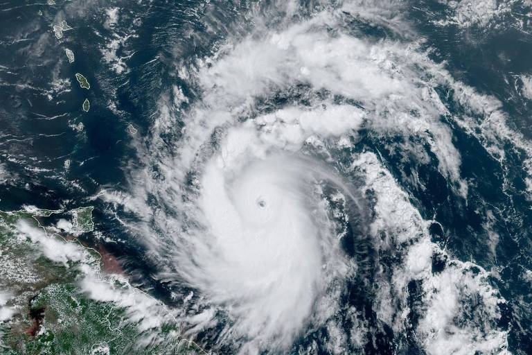 Imagem de satélite mostra um furacão no oceano. O furacão tem uma forma espiral com um olho bem definido no centro. As nuvens densas e brancas se estendem em espiral a partir do olho do furacão. A terra é visível no canto inferior esquerdo da imagem, enquanto o resto da imagem é dominado pelo oceano e pelo furacão.